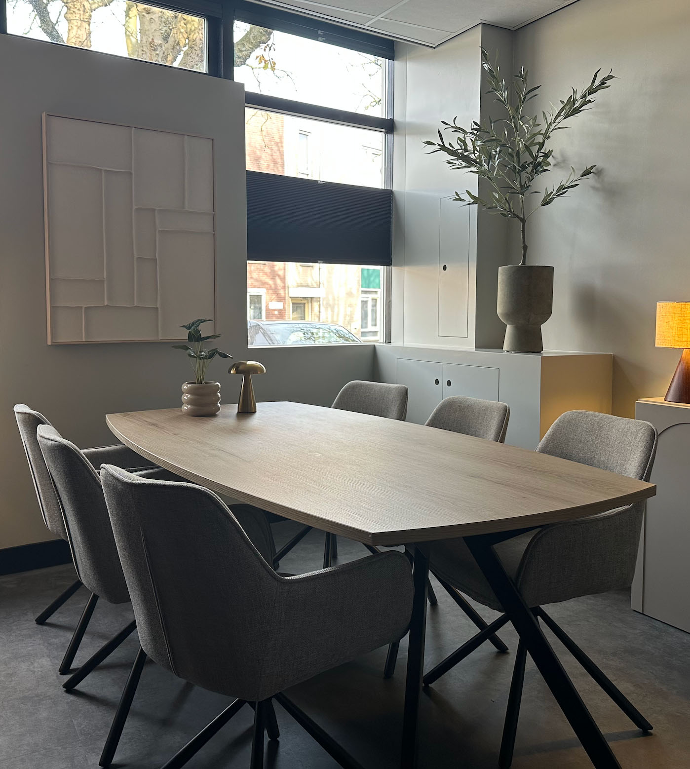 Moderne vergaderruimte met houten tafel, grijze stoelen, planten en grote ramen met jaloezieën