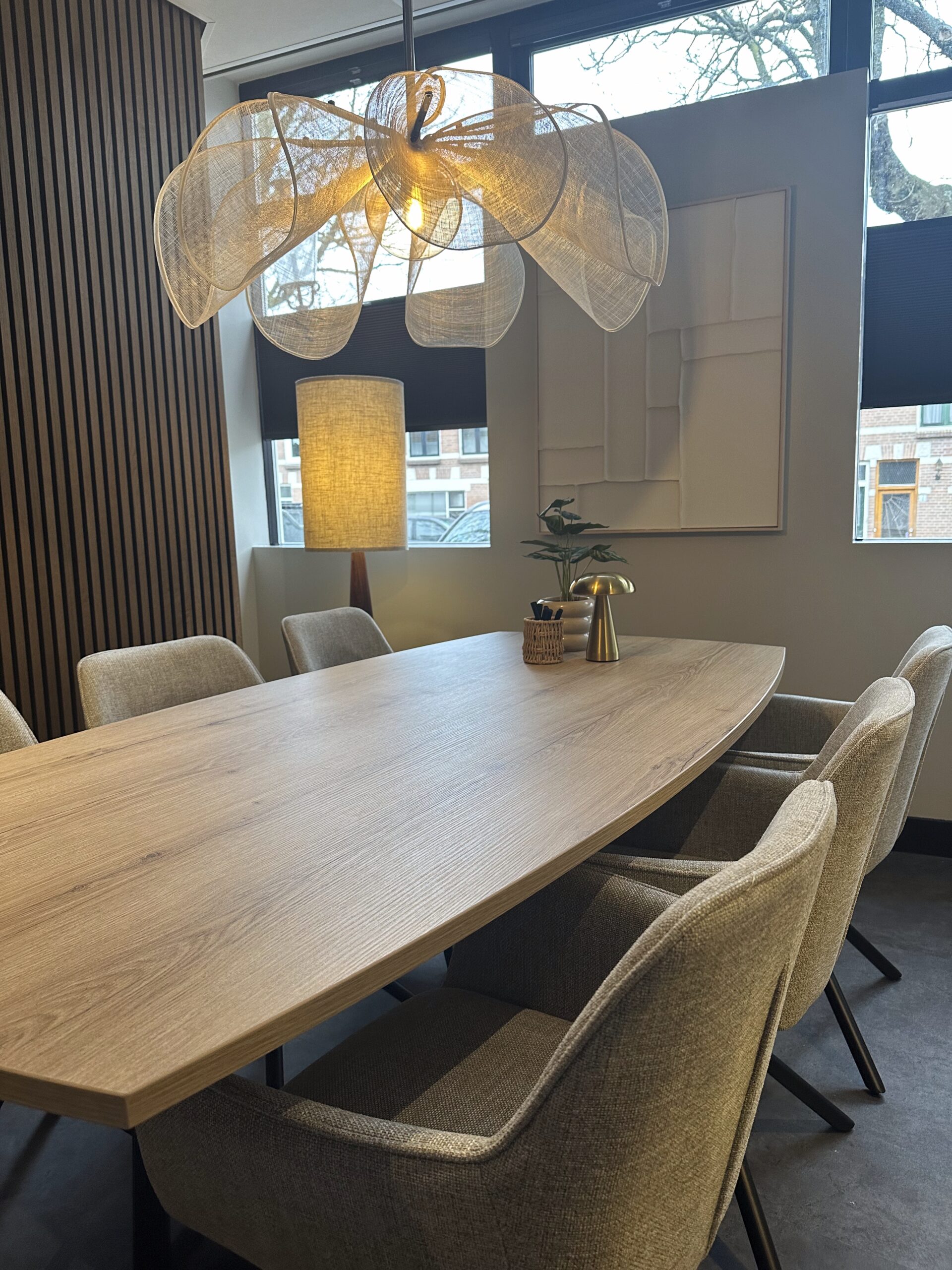 Vergaderruimte met houten tafel, beige stoelen en een hanglamp met gouden lampenkappen